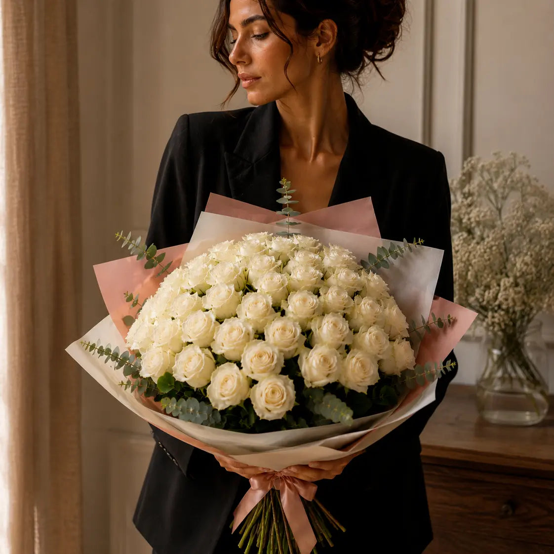 Modelo segurando um ramo premium de rosas brancas com luz natural, papel elegante, eucalipto e laço rosa.