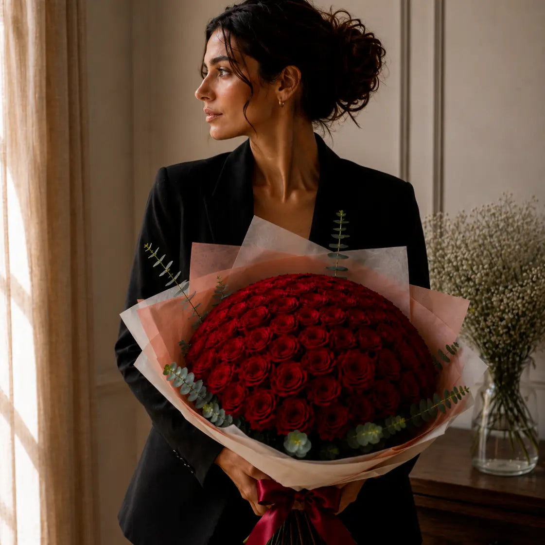 Modelo junto à janela segurando um ramo premium de rosas vermelhas com papel elegante, eucalipto e laço vinho tinto.