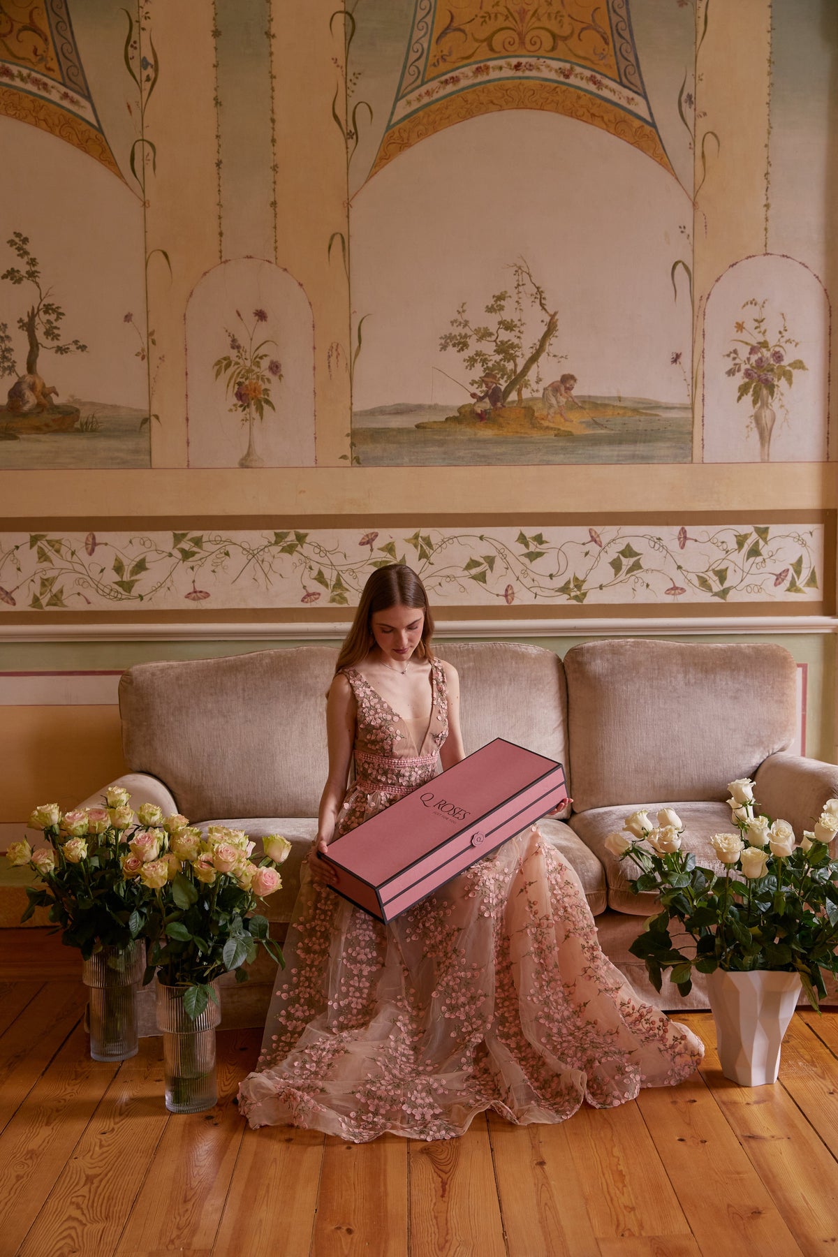 Mulher com uma caixa QRoses cor-de-rosa, rodeada de ramos de rosas numa sala.