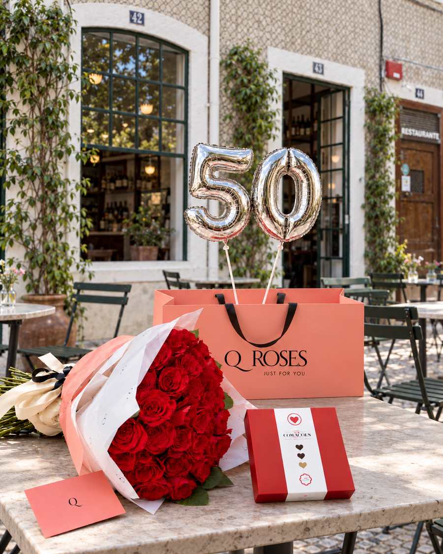 Presente QRoses Grand Celebration com ramo de rosas vermelhas, chocolates, envelope e balões prateados com o número 50, sobre uma mesa junto a uma fachada de azulejos.