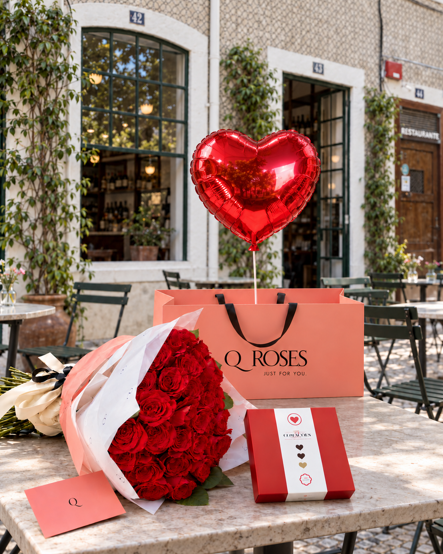 Presente romântico QRoses Grand Celebration com rosas vermelhas, chocolates, envelope e balão vermelho em forma de coração, sobre uma mesa junto a uma fachada de azulejos.