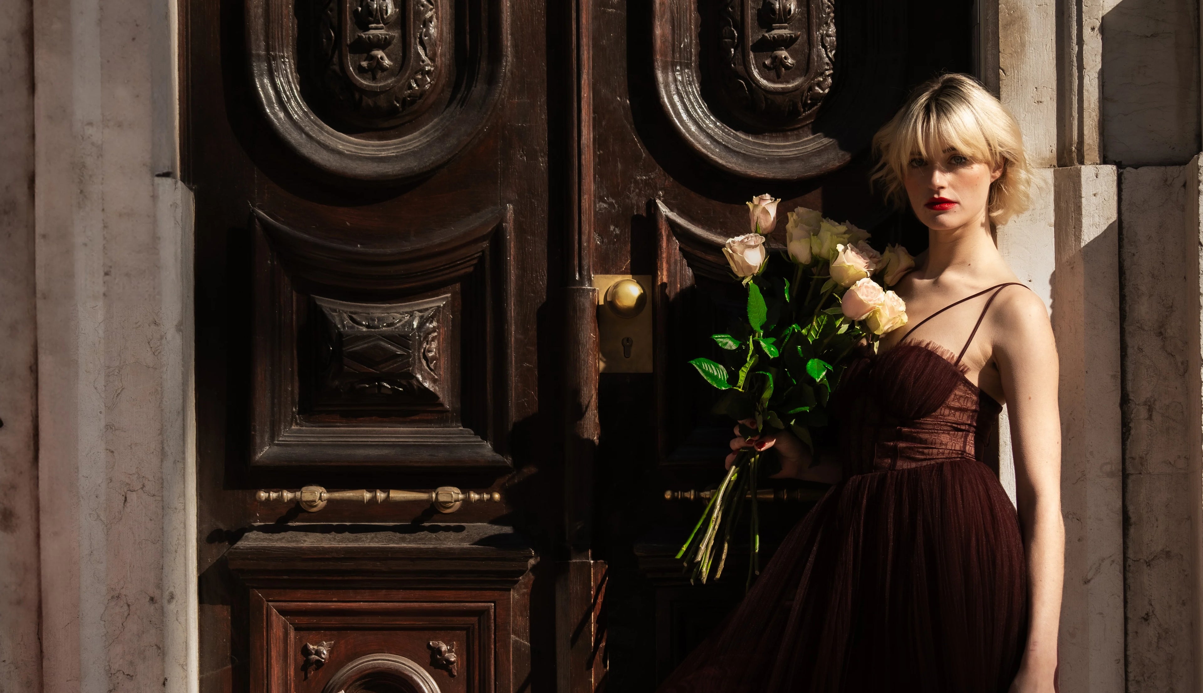 Elegant woman in a burgundy dress holding a bouquet of premium blush roses outside a historic wooden doorway in Lisbon, showcasing luxury floral gifting by QROSES.