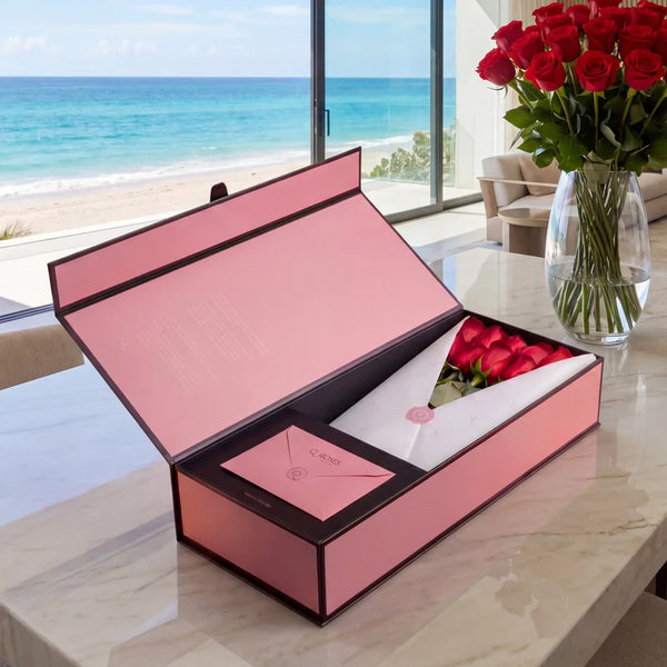 Caixa Signature Box QRoses com rosas vermelhas premium, cartão QR e embalagem rosa elegante sobre mesa de mármore com vista para o mar.