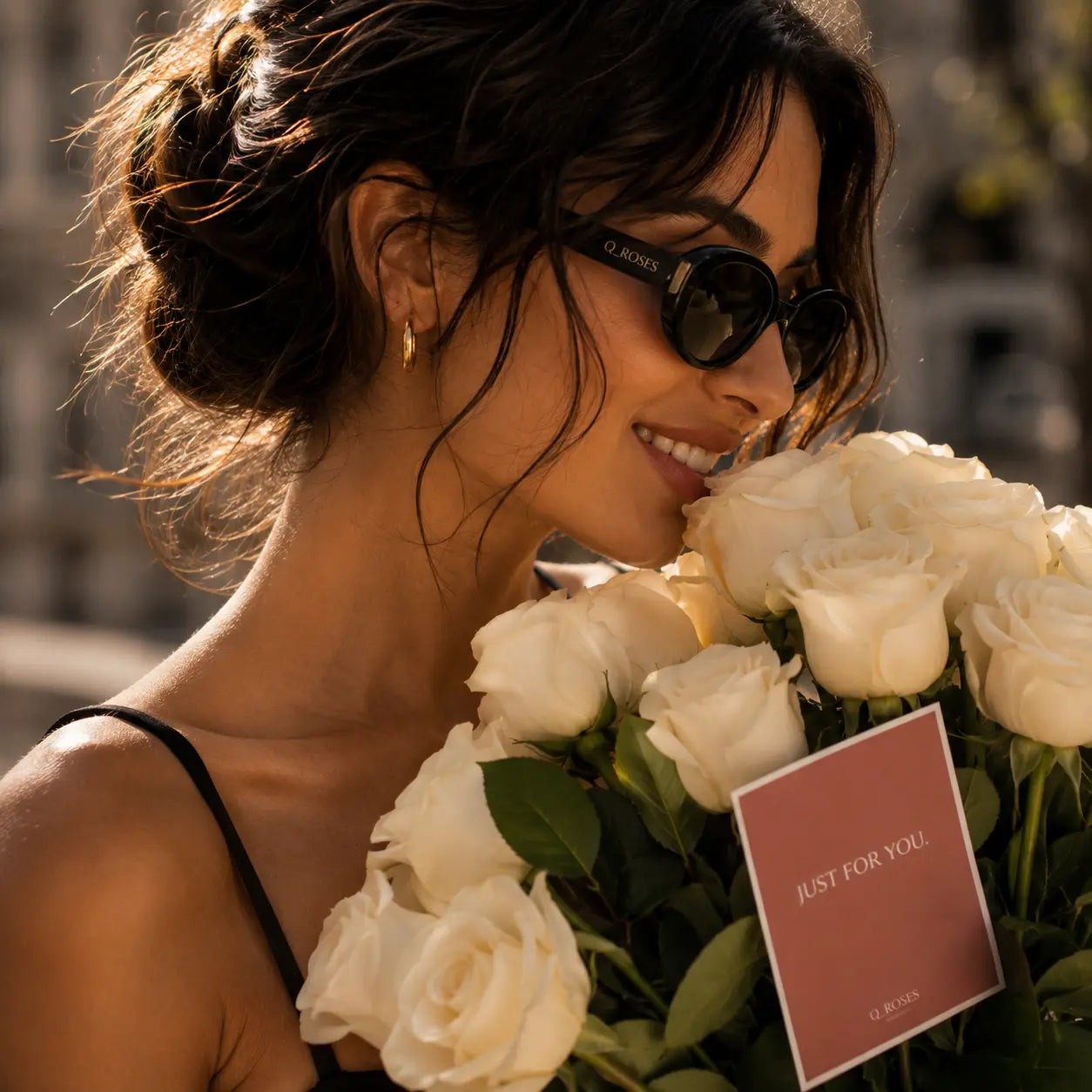 Modelo elegante com óculos escuros a sorrir enquanto segura bouquet de rosas brancas premium QRoses com cartão personalizado.