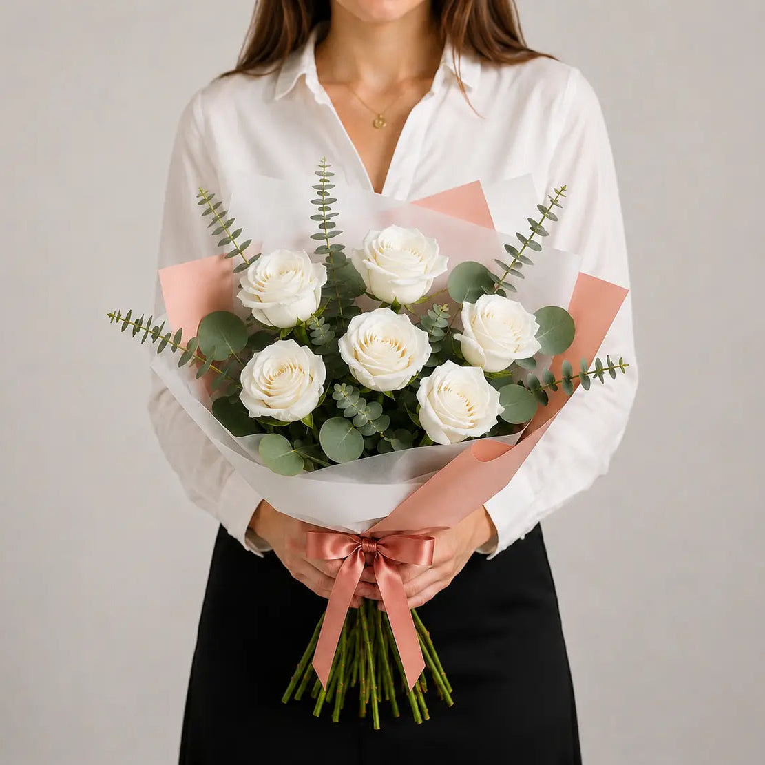 Mulher a segurar um Ramo Essencial de 5 ou 6 rosas brancas, envolvido em papel artístico branco e rosa, com eucalipto e laço acetinado rosa.