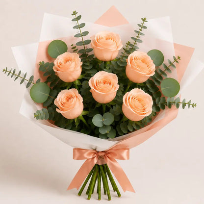 Ramo Essencial de Rosas Cor-de-Pêssego da QRoses, cuidadosamente embalado com papel artístico branco e rosa, folhagem de eucalipto e laço acetinado, fotografado sobre fundo claro.