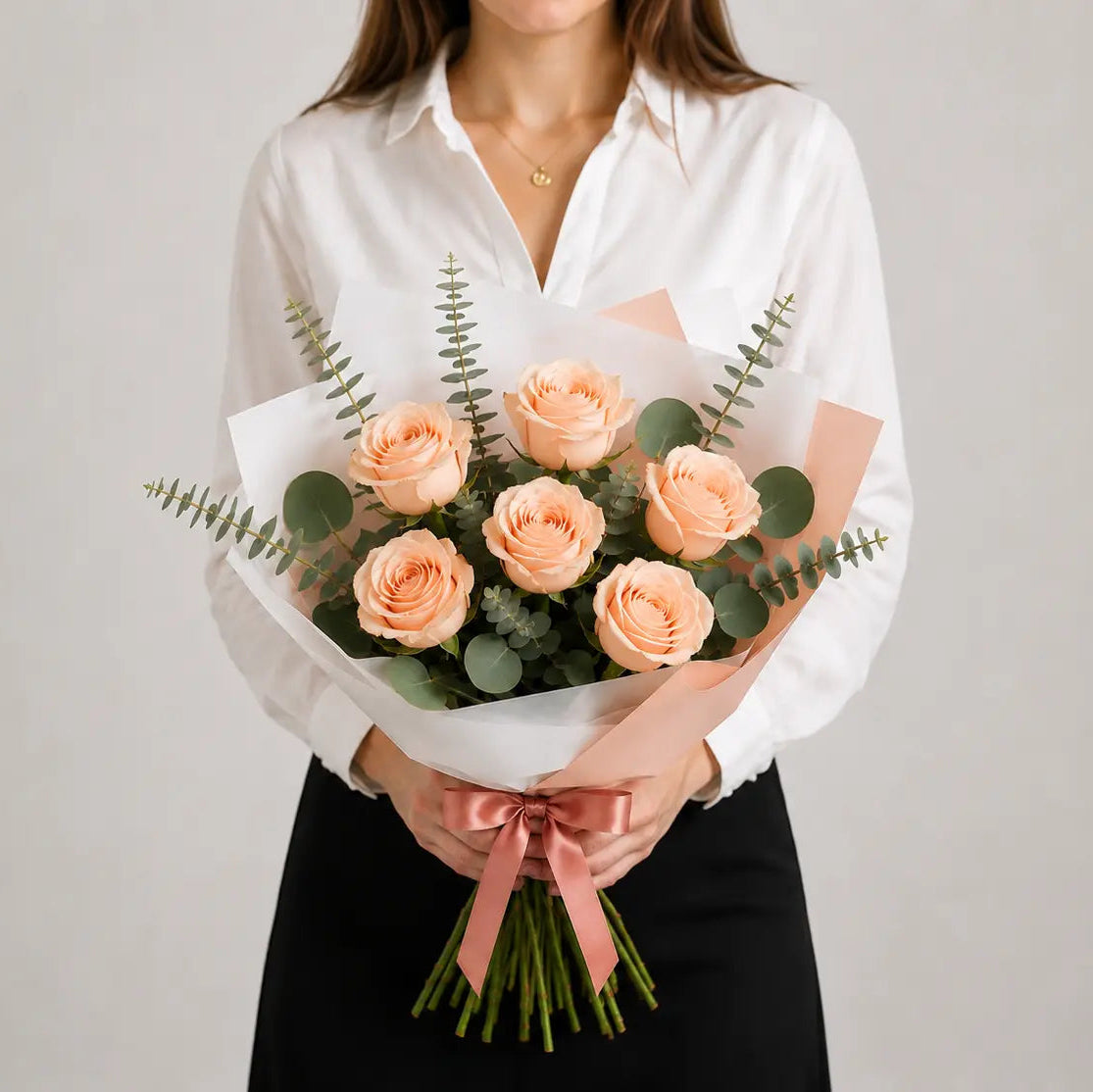Modelo a segurar um Ramo Essencial de Rosas Cor-de-Pêssego da QRoses, cuidadosamente embalado com papel artístico branco e rosa, folhagem de eucalipto e laço acetinado.