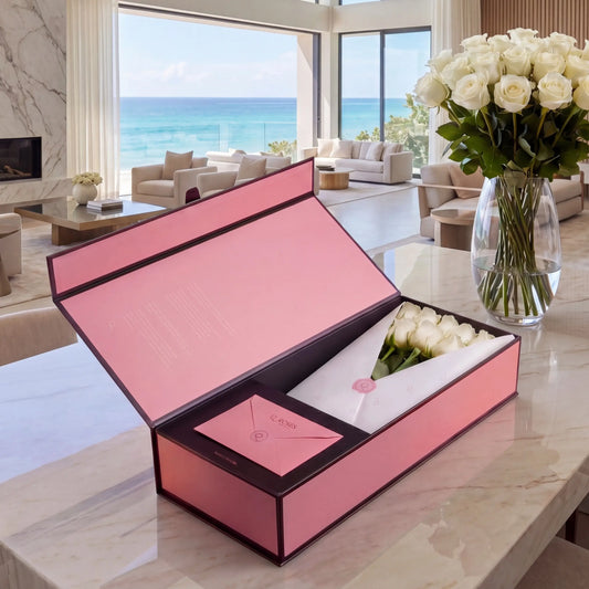 Caixa Signature Box QRoses com rosas brancas premium, cartão QR e embalagem rosa elegante sobre mesa de mármore num ambiente de luxo à beira-mar.