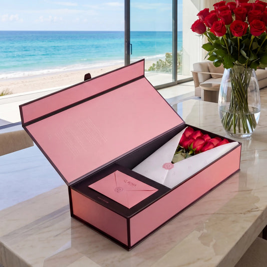 Caixa Signature Box QRoses com rosas vermelhas premium, cartão QR e embalagem rosa elegante sobre mesa de mármore com vista para o mar.