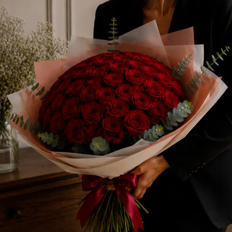 Detalhe de modelo segurando um ramo premium de rosas vermelhas com papel elegante, eucalipto e laço vinho tinto.