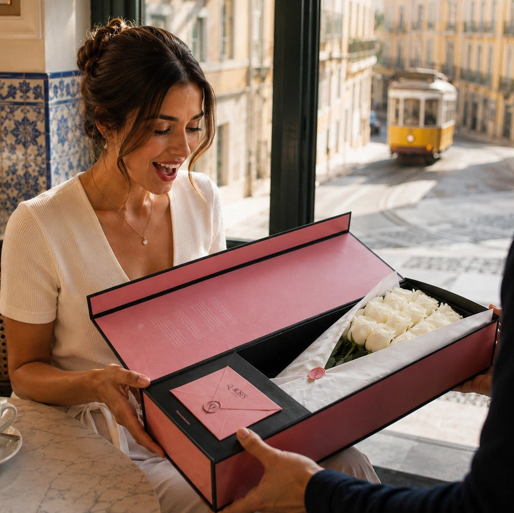 Elegant woman receiving a QROSES luxury gift box with premium white roses in a Lisbon café, featuring the signature pink and black presentation box with QR Card and premium wrapping.