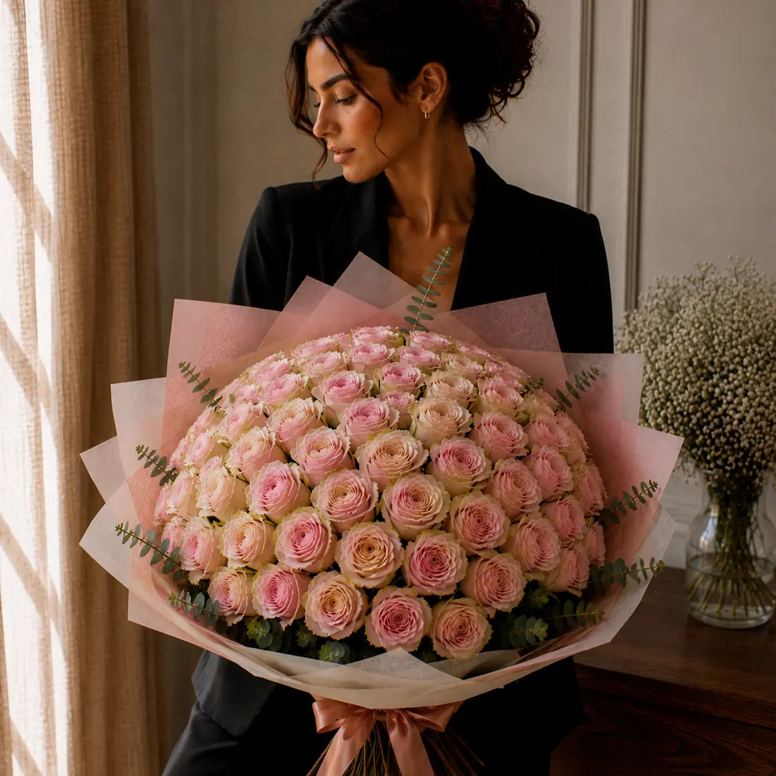 Modelo segurando um ramo premium de rosas rosadas com bordes esverdeados num interior elegante, com papel sofisticado, eucalipto e laço rosa.