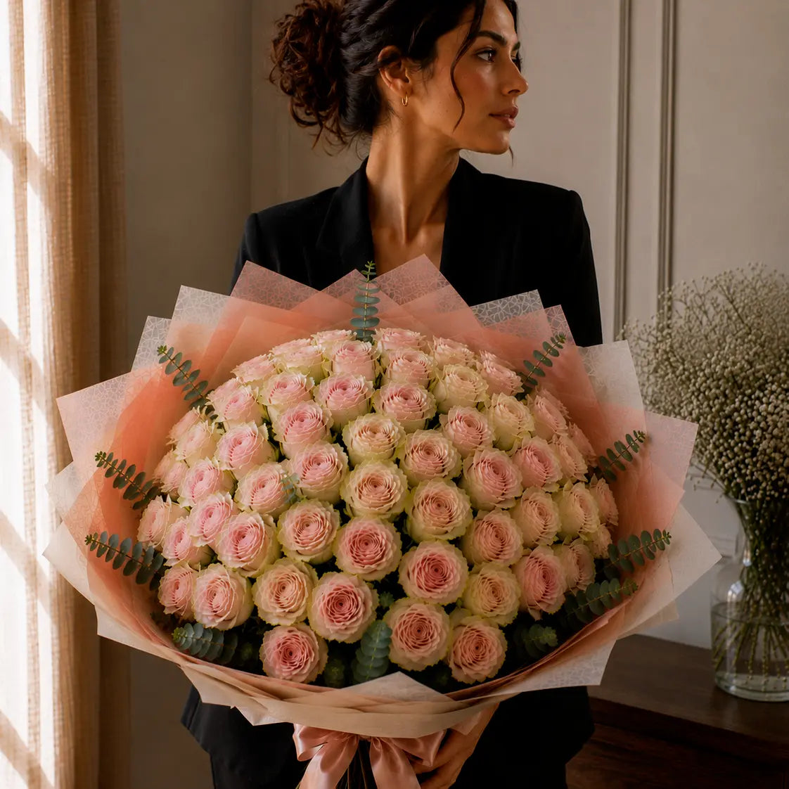 Modelo segurando um ramo premium de rosas rosadas com bordes esverdeados junto à janela, com papel elegante, eucalipto e laço rosa.