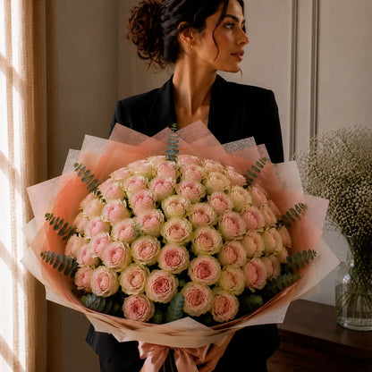 Modelo segurando um ramo premium de rosas rosadas com bordes esverdeados junto à janela, com papel elegante, eucalipto e laço rosa.