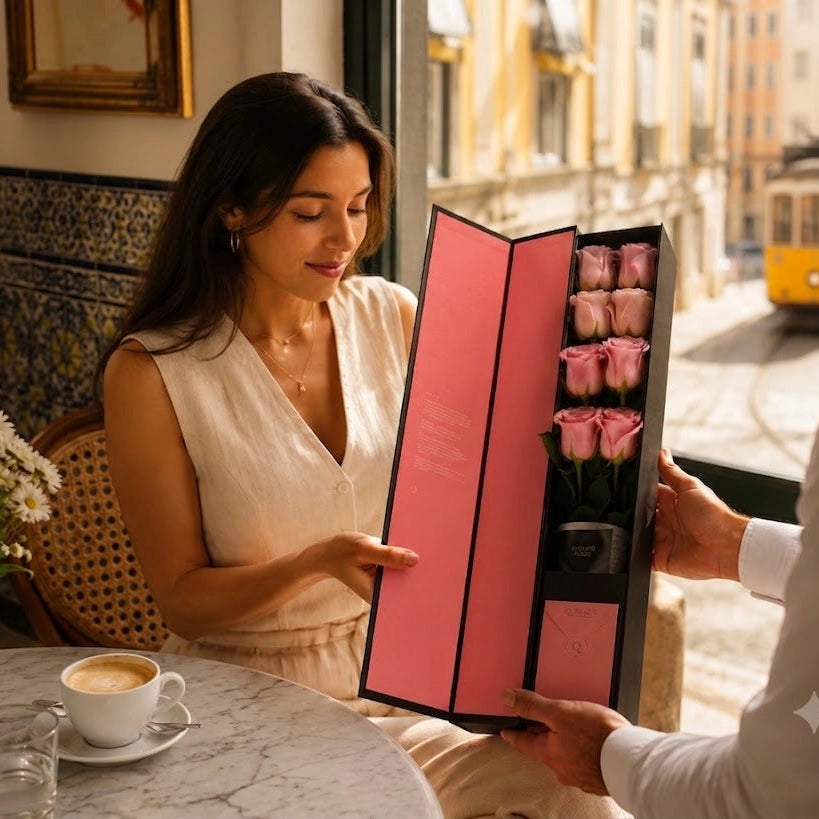 Elegant woman receiving a QROSES FLUTE gift box with eight premium soft pink roses featuring delicate green outer petals inside a sunlit Lisbon café, luxury floral gift experience.
