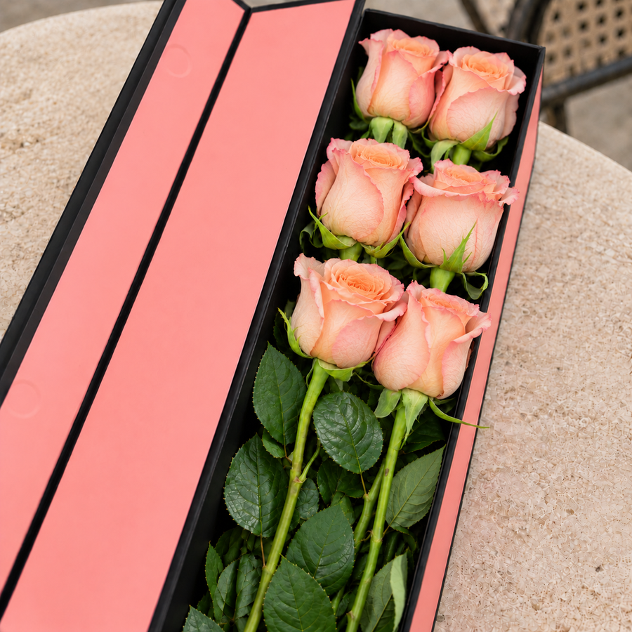 Caixa Flute QRoses aberta, em rosa e preto, com seis rosas naturais cor de pêssego Shimmer e folhagem verde sobre uma mesa de pedra.