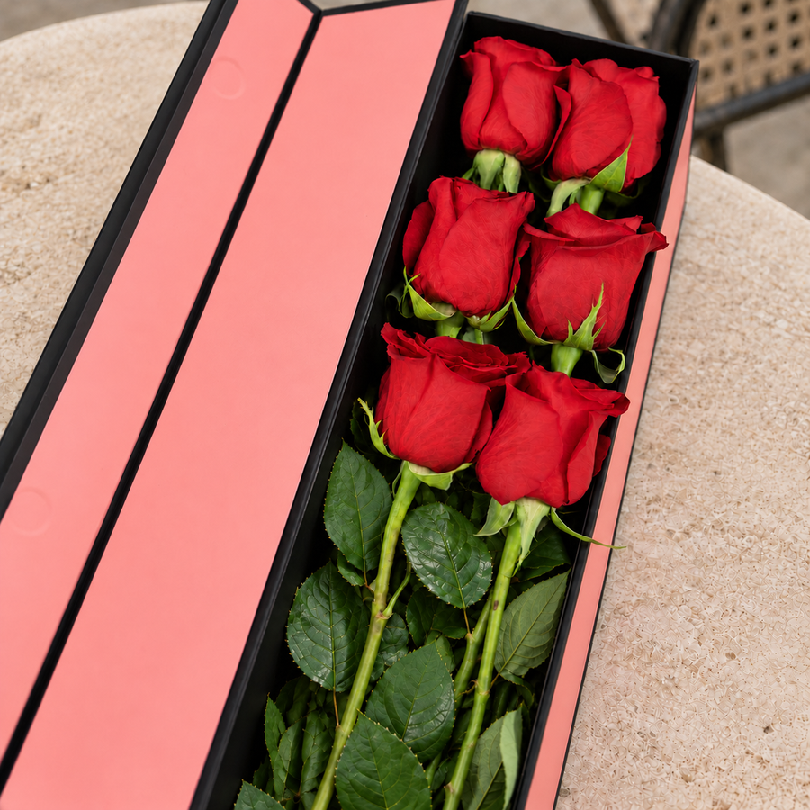 Caixa Flute QRoses aberta, em rosa e preto, com seis rosas vermelhas naturais e folhagem verde sobre uma mesa de pedra.
