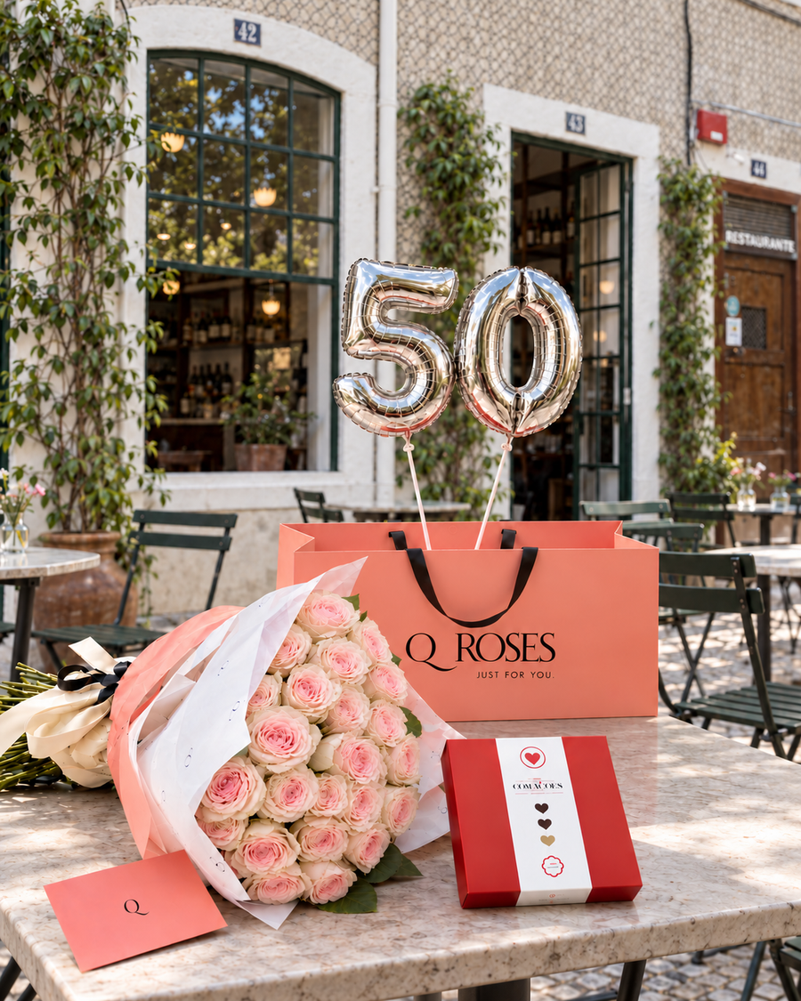Presente de aniversário QRoses Grand Celebration com rosas cor-de-rosa Fruttetto, chocolates, envelope e balões prateados com o número 50, sobre uma mesa junto a uma fachada de azulejos em Lisboa.