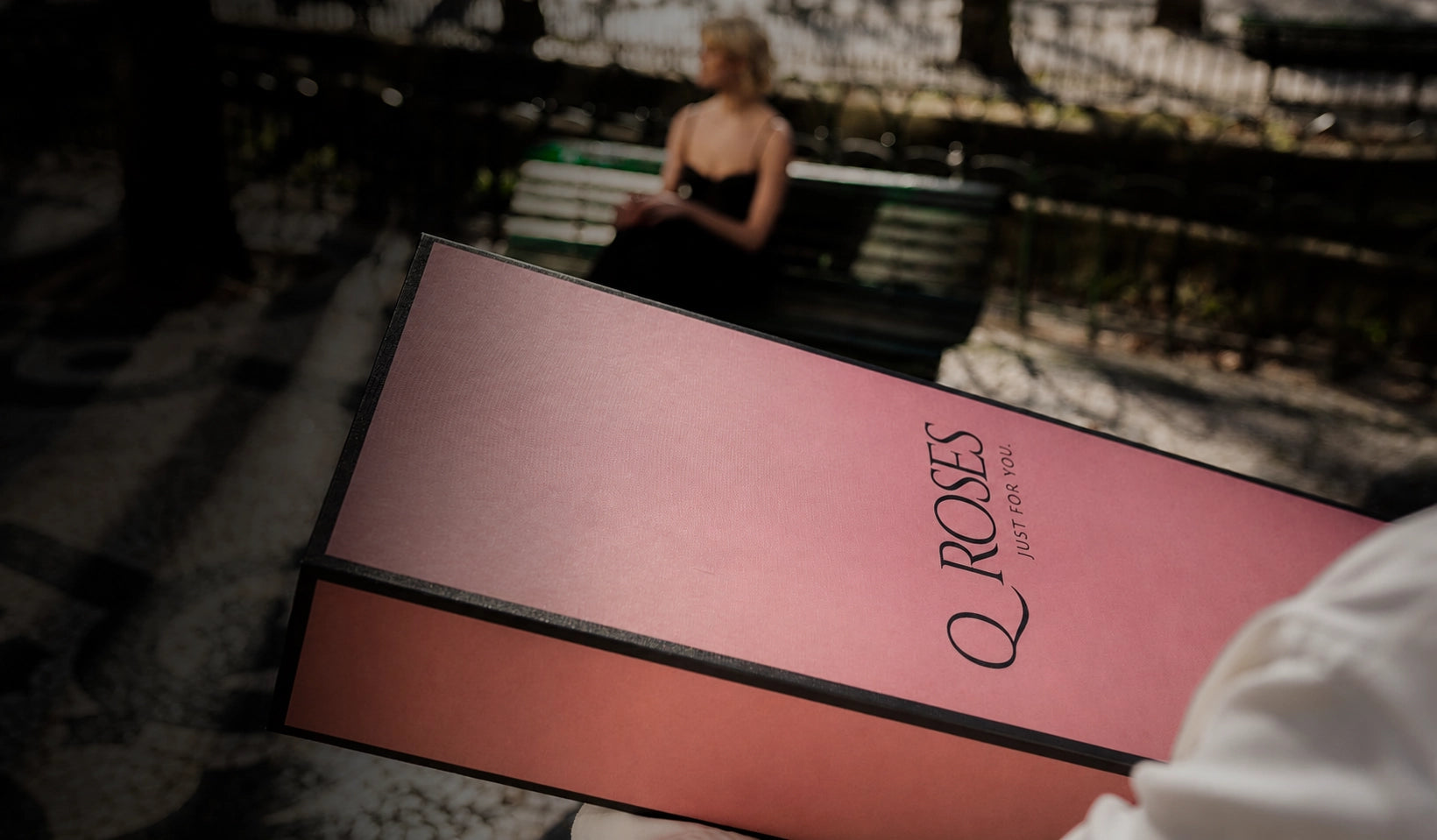 Luxury QRoses Signature Box being delivered in a park, with the elegant pink gift box in focus and the recipient waiting in the background.