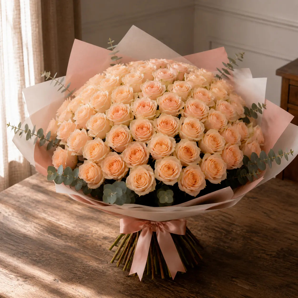 Ramo premium de rosas em tom pêssego claro sobre mesa de madeira, com papel elegante, eucalipto e laço rosa.
