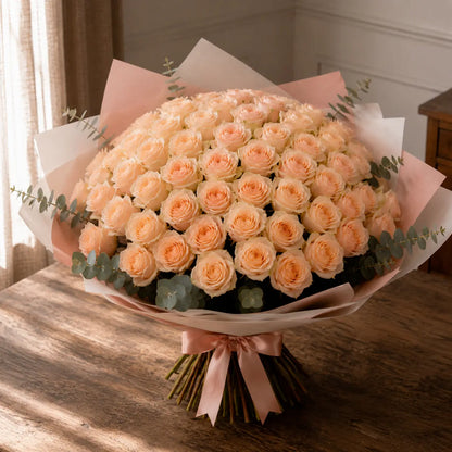 Ramo premium de rosas em tom pêssego claro sobre mesa de madeira, com papel elegante, eucalipto e laço rosa.