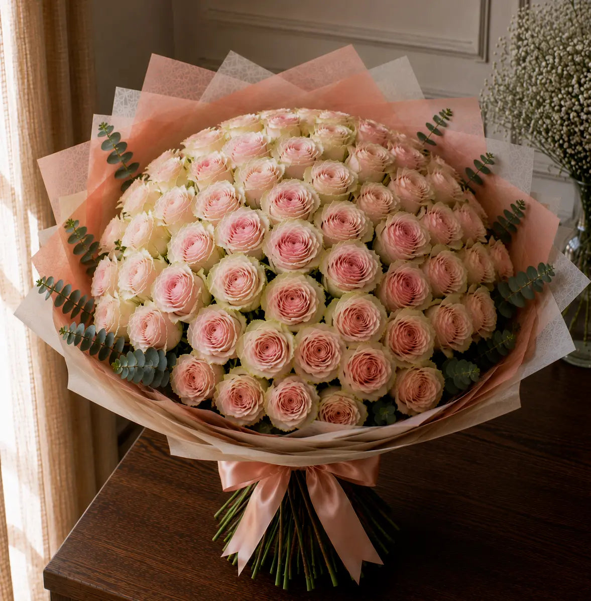 Ramo premium de rosas rosadas com bordes esverdeados sobre mesa de madeira, com papel elegante, eucalipto e laço rosa.