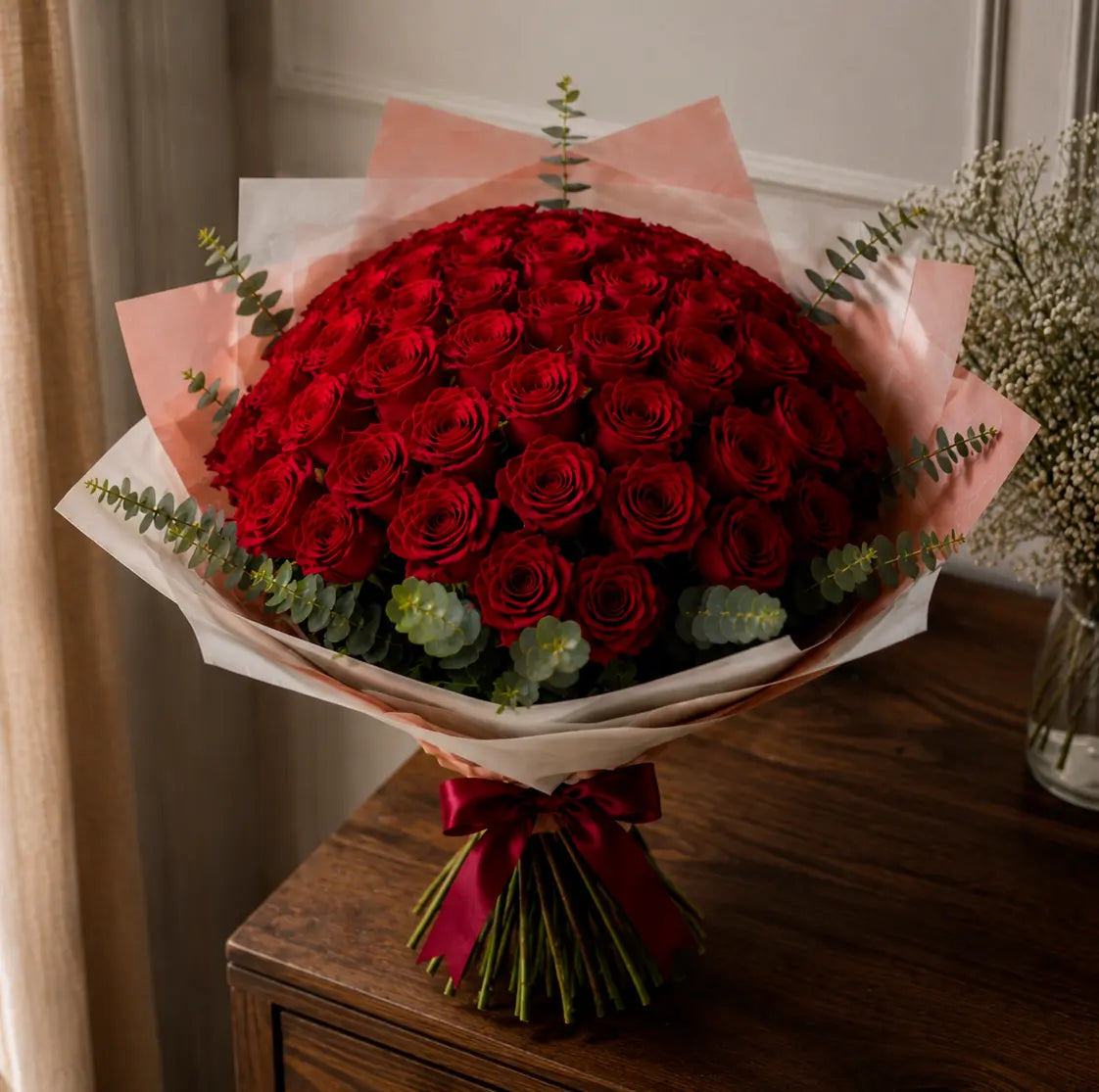 Ramo premium de rosas vermelhas sobre mesa de madeira, com papel elegante, folhagem de eucalipto e laço vinho tinto.