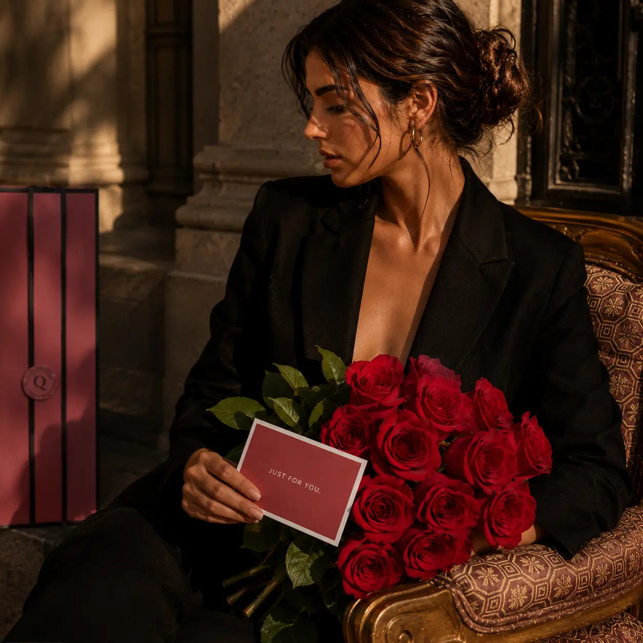 Mulher elegante segurando bouquet de rosas vermelhas premium QRoses com cartão, sentada num ambiente sofisticado e acolhedor.