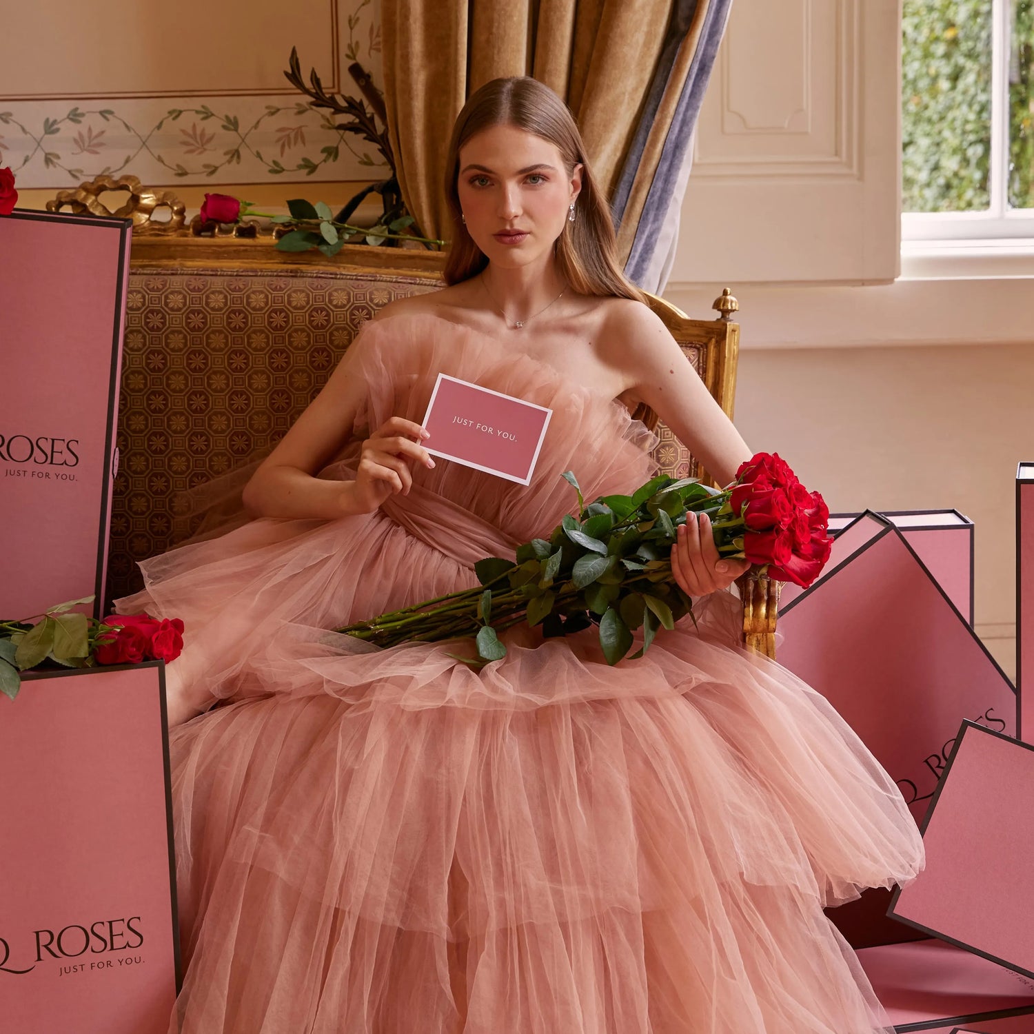 Elegant woman in a blush couture gown holding premium red roses with QRoses Signature Boxes in a luxury interior, showcasing exclusive floral gifting.